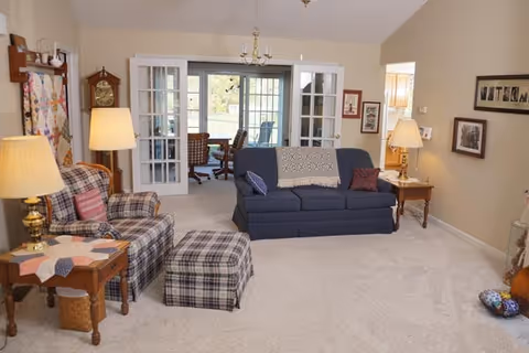 A cozy living room with beige walls and carpeted floor featuring a blue sofa with pillows and a throw blanket, a plaid armchair with matching ottoman, two wooden side tables with lamps, a grandfather clock, and framed artwork on the walls. French doors lead to a sunroom with additional seating and a view of the outdoors.