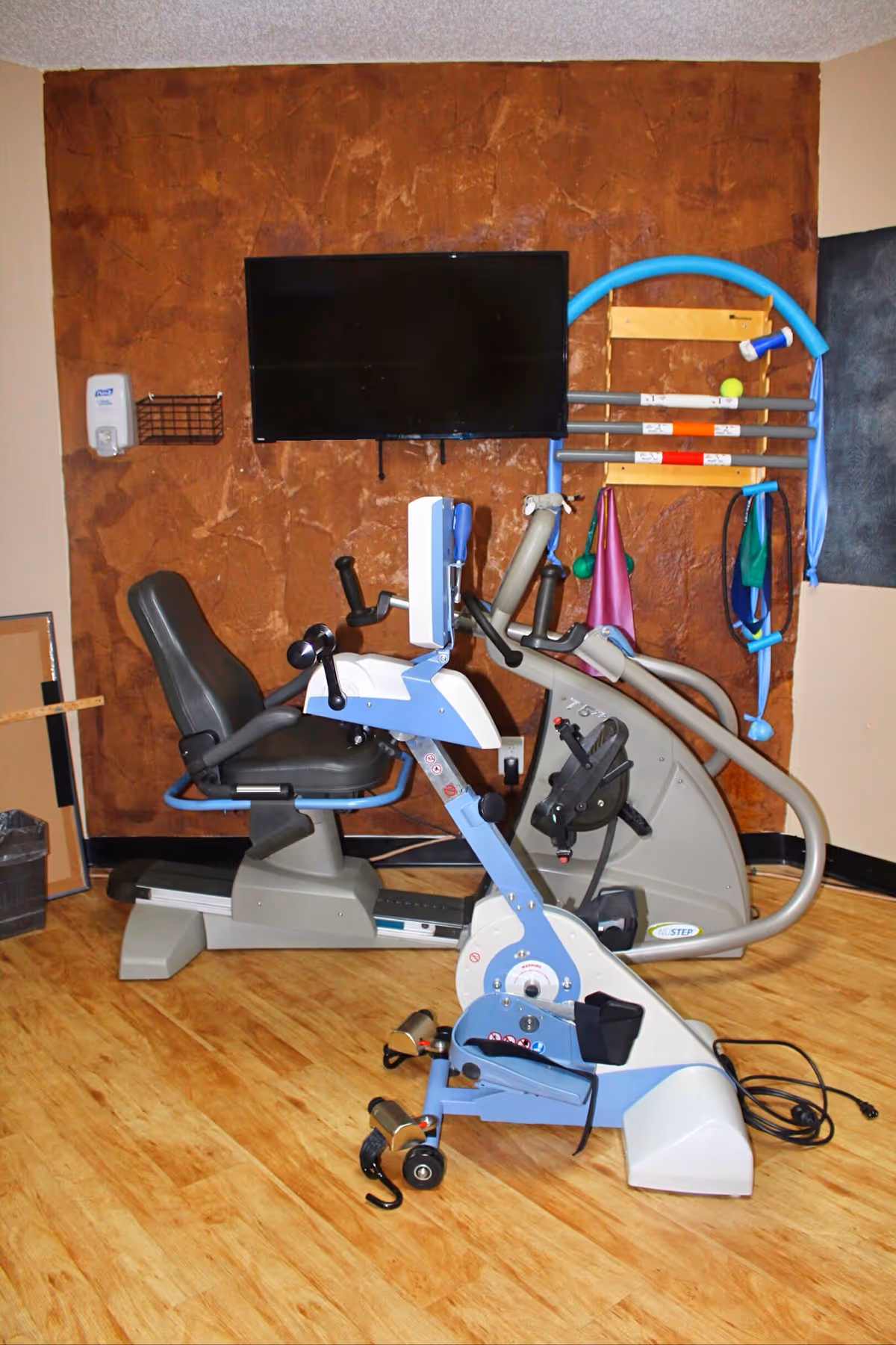 A small exercise room with a recumbent stationary bike and a NuStep exercise machine on a wooden floor. A flat-screen TV is mounted on a textured brown wall. Various exercise accessories, including resistance bands and a blue pool noodle, hang on a rack attached to the wall. A hand sanitizer dispenser is mounted on the left side of the wall.