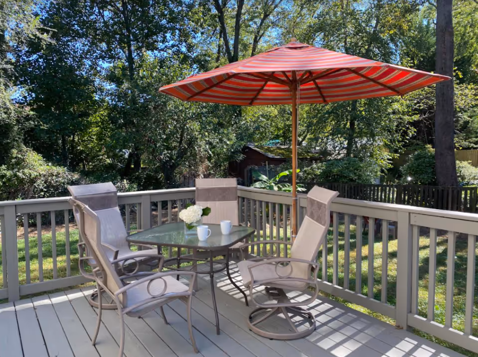 Outdoor patio area with a glass-top table, four beige mesh chairs, a red and white striped umbrella, and a small vase with white flowers. The patio overlooks a green yard with trees and bushes in the background.