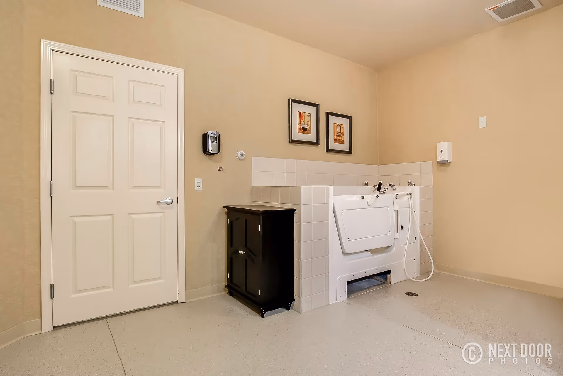 A beige-colored room with a white door on the left, a black cabinet next to a white walk-in bathtub with a door and hand-held shower. Two framed pictures hang on the wall above the bathtub. There is a soap dispenser mounted on the wall near the door and another dispenser on the adjacent wall.