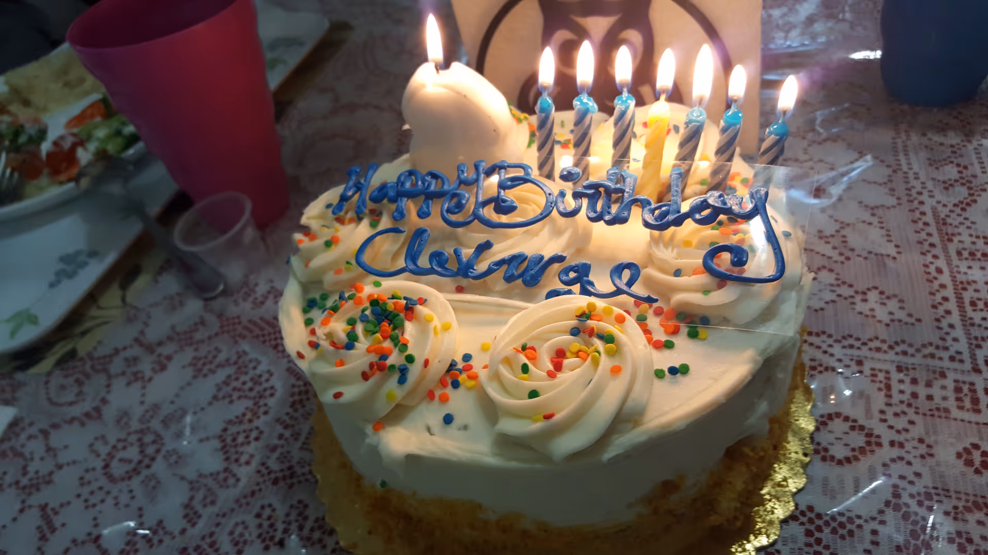 A birthday cake with white frosting, colorful sprinkles, and lit candles on top. The cake has a message written in blue icing that says 'Happy Birthday Clarence'. The cake is placed on a table with a lace tablecloth, and there are plates with food and a pink cup in the background.