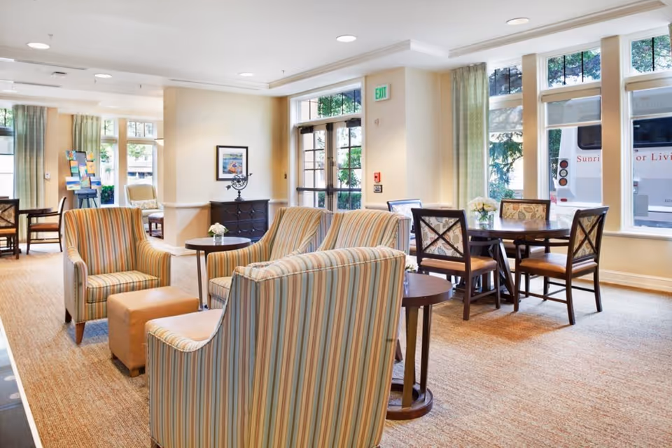 A bright and spacious common area in a senior living facility with striped upholstered armchairs, a small round table with a flower vase, and a dining table with four chairs near large windows. Outside the windows, a Sunrise Senior Living shuttle bus is visible.