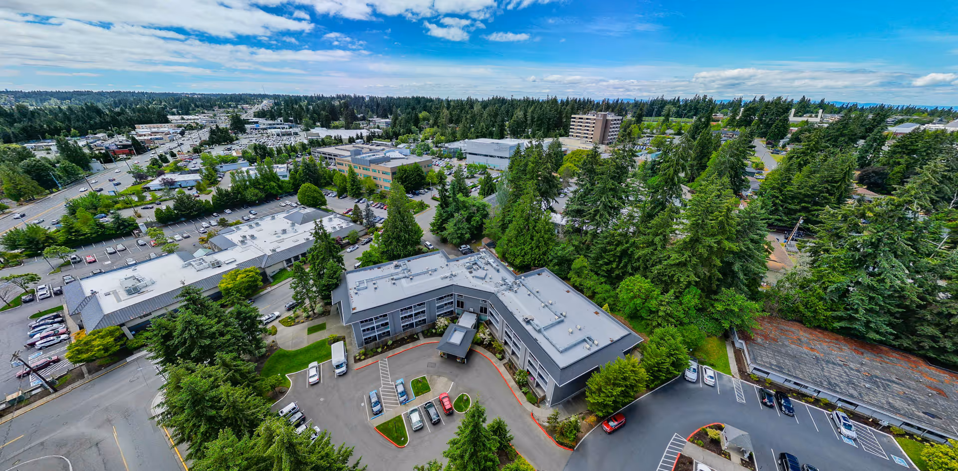 Aerial view of Aegis of Edmonds - Assisted Living facility surrounded by trees and parking lots, with a clear blue sky and scattered clouds in the background.