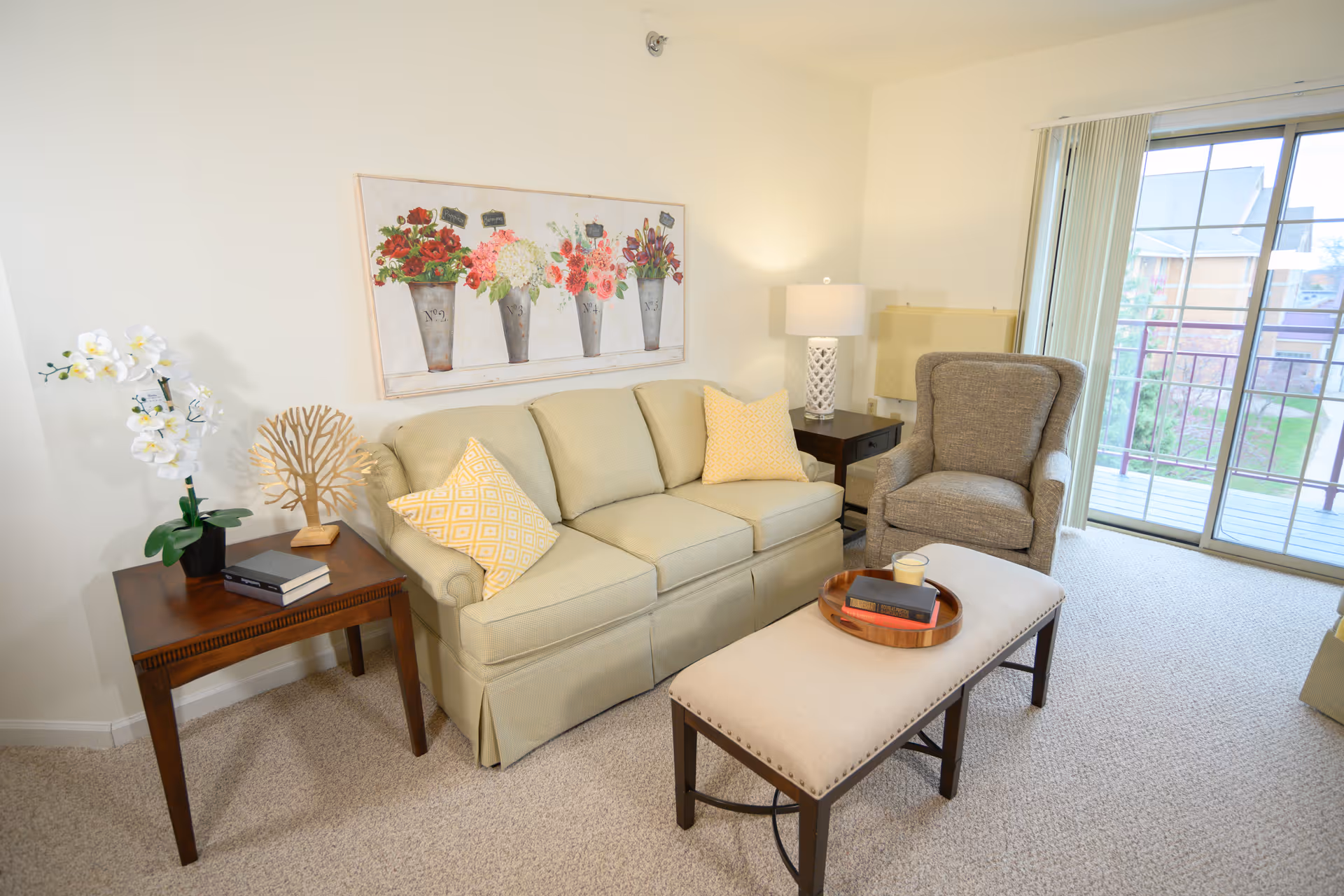 A cozy living room with a beige sofa adorned with two yellow patterned pillows, a gray armchair, and a cushioned bench with a wooden tray holding books and a candle. There is a wooden side table with a white orchid plant and decorative item, a table lamp on a dark wooden side table, and a large floral painting on the wall. Sliding glass doors lead to a balcony with a view of neighboring buildings and greenery.