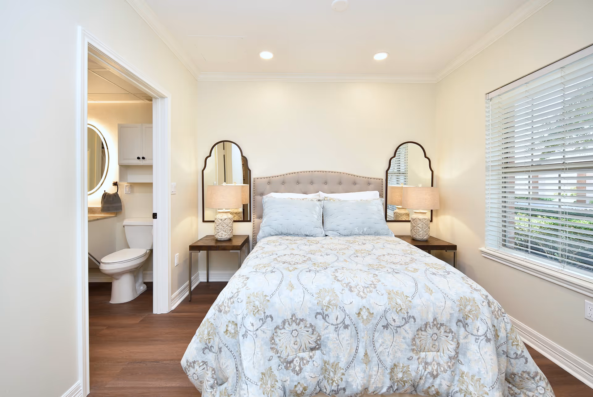 A bright bedroom with a neatly made bed featuring a patterned comforter and two blue pillows. On either side of the bed are wooden nightstands with matching lamps and decorative mirrors above them. To the left, there is an open door leading to a bathroom with a visible toilet and a round illuminated mirror above the sink. A window with white blinds is on the right wall, allowing natural light into the room.