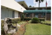 Outdoor garden area at Park Ridge Nursing and Rehabilitation with a small rock water feature, green grass, shrubs, and palm trees near a building with windows and an American flag on a flagpole.