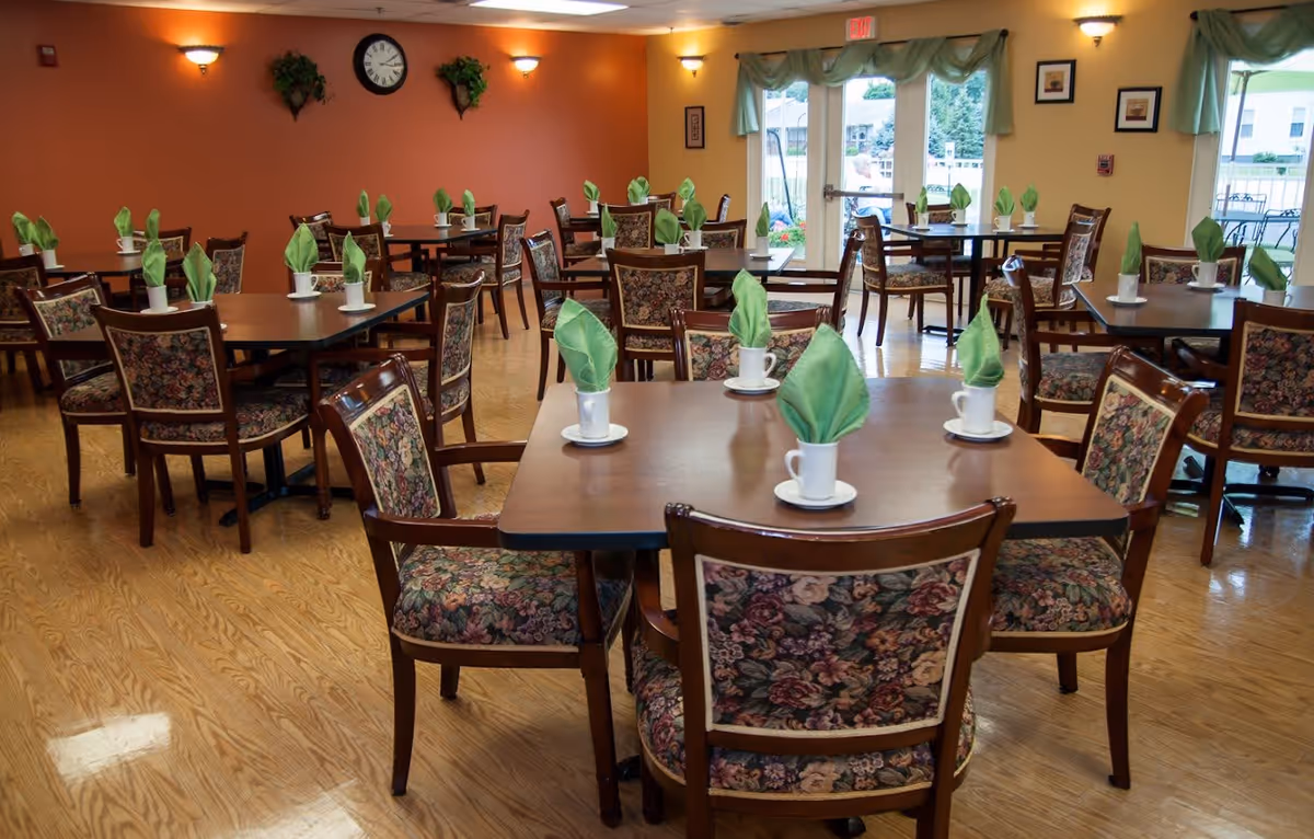 A dining room with multiple wooden tables and floral upholstered chairs. Each table has white cups with green folded napkins inside. The room has wooden flooring, orange and yellow walls, wall-mounted lights, a clock, and large windows with green curtains letting in natural light.