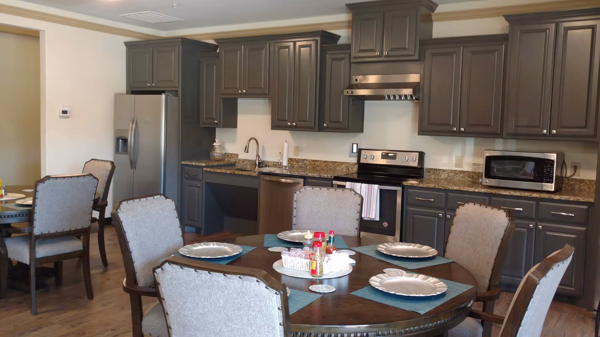 A kitchen and dining area in an assisted living community featuring dark gray cabinets, stainless steel appliances including a refrigerator, stove, dishwasher, and microwave. A round wooden dining table is set with plates, placemats, and condiments, surrounded by upholstered chairs.