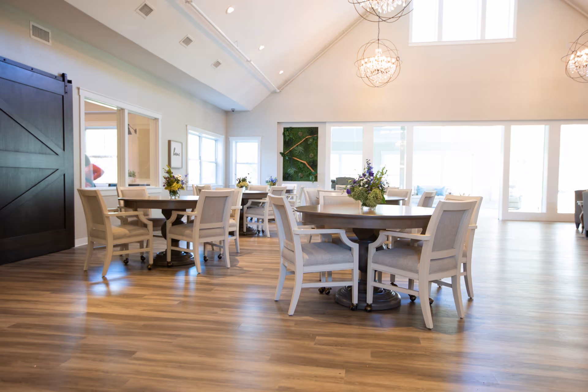 Bright, airy communal dining room with round tables, upholstered chairs, floral centerpieces, and large windows.