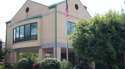 Two-story beige building with large windows, green trim, flagpole, and surrounding trees.