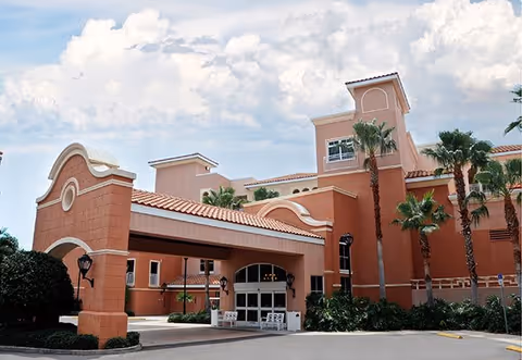 Exterior view of Westminster Shores facility featuring a large entrance with a covered driveway, terracotta-colored walls, palm trees, and a partly cloudy sky.