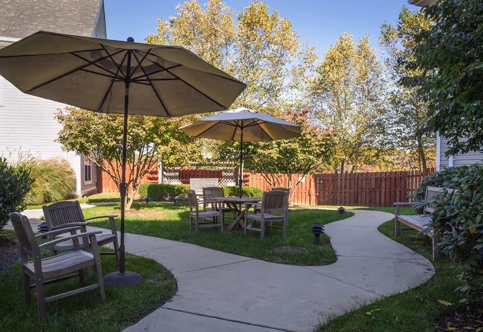 Outdoor seating area in a garden with wooden chairs and tables under large beige umbrellas, surrounded by green grass, trees, and a wooden fence in the background.