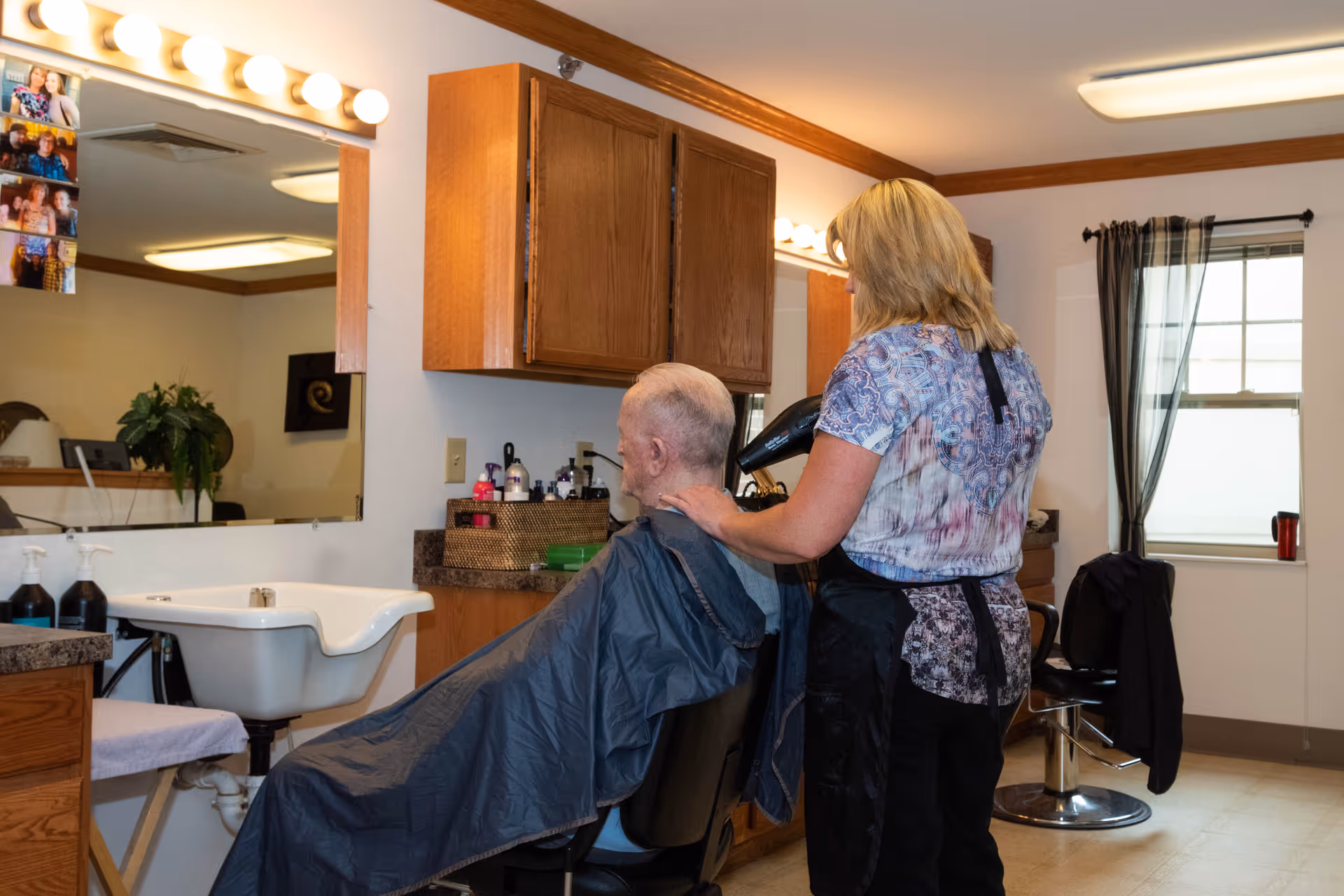 A hairdresser blow-drying the hair of an elderly man seated in a salon chair inside a well-lit room with wooden cabinets, a large mirror, and a wash basin. The room has a window with sheer curtains and various hair care products on the counter.