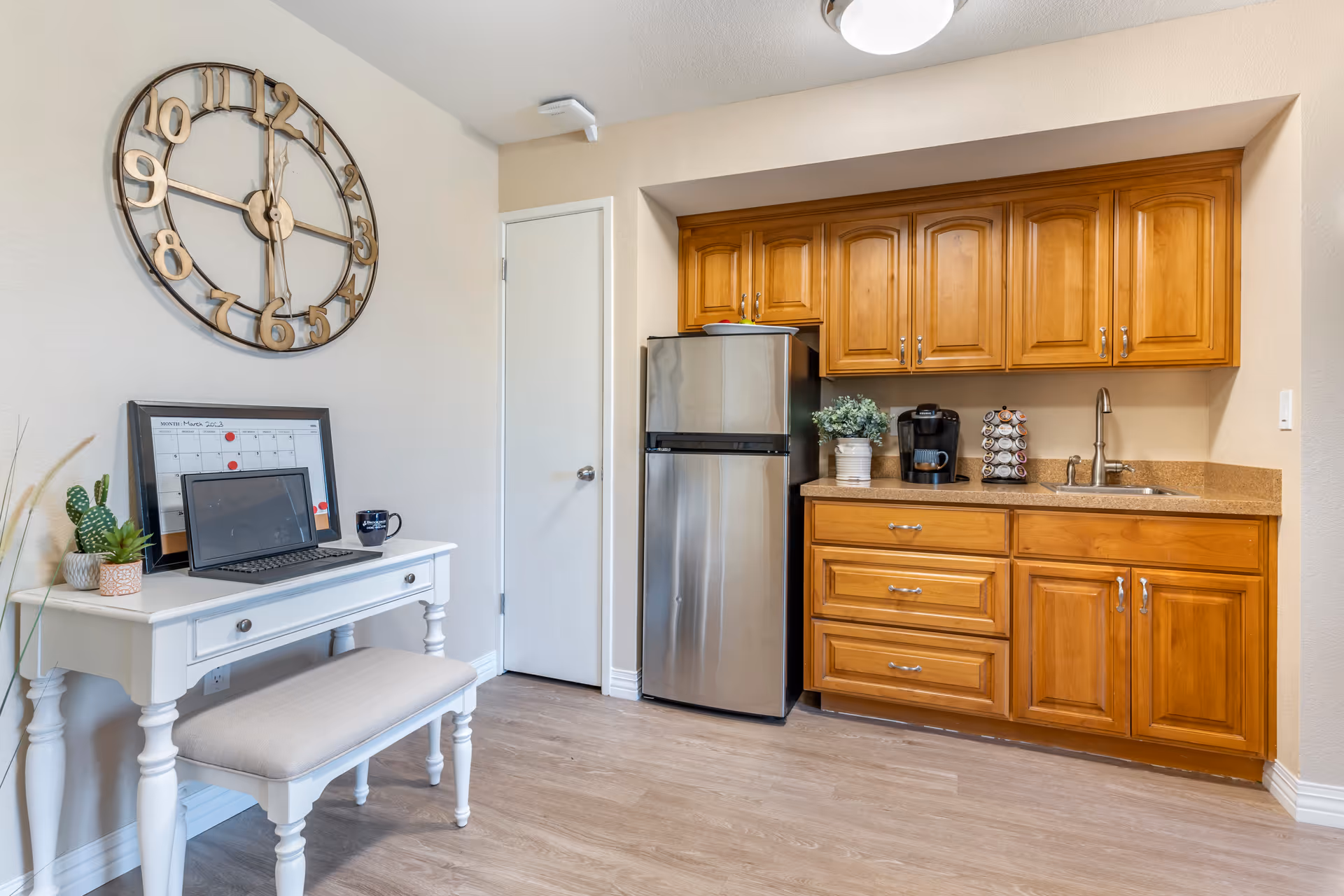 A small kitchen area with wooden cabinets, a stainless steel refrigerator, a coffee maker with a pod holder, and a sink. To the left, there is a white desk with a cushioned bench, a laptop, a calendar, a coffee mug, and small potted plants. A large decorative wall clock is mounted above the desk on a beige wall.