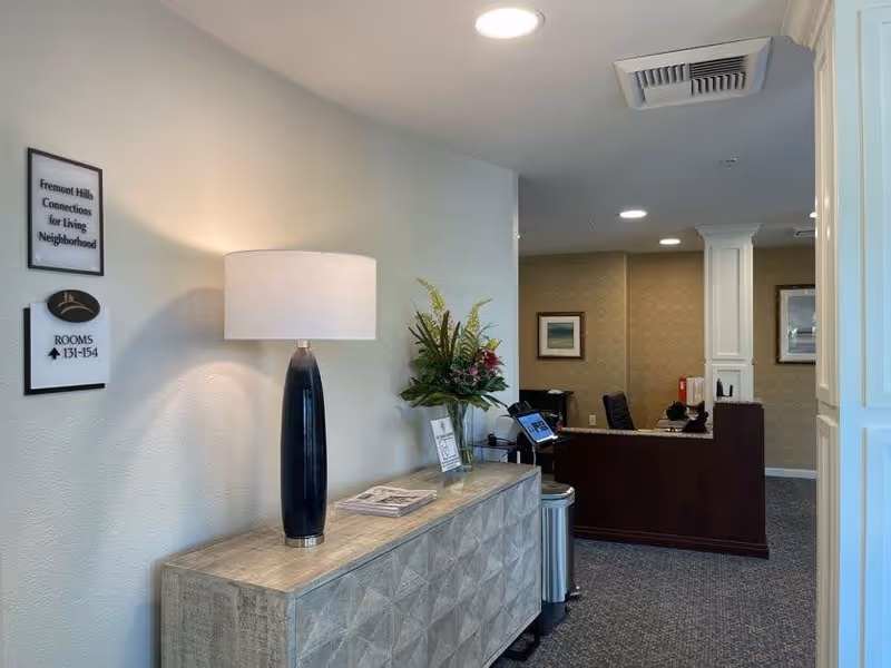 Interior view of a reception area in Fremont Hills facility with a wooden sideboard holding a lamp, a flower arrangement, and some papers. A sign on the wall indicates rooms 131-154 are in the direction shown. In the background, there is a reception desk with a computer and office chair, and framed artwork on the walls.