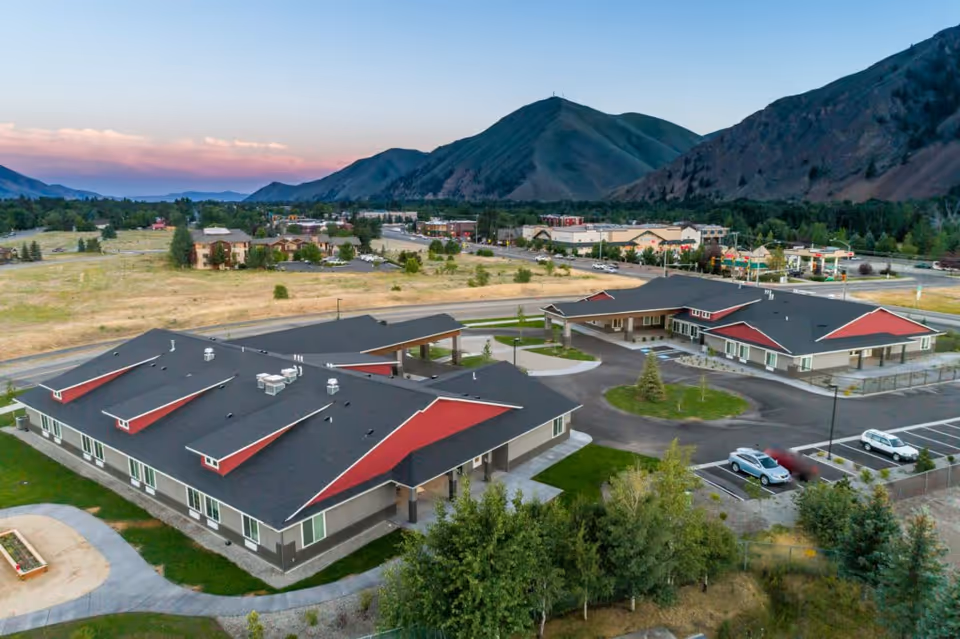 Aerial view of Silvercreek Senior Living facility with two large buildings featuring dark roofs and red accents, surrounded by parking lots, trees, and open fields with mountains in the background under a clear sky at sunset.