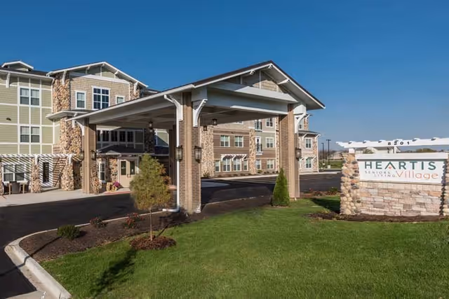 Exterior view of Heartis Village of Peoria senior living facility showing the main entrance with a covered driveway, landscaped lawn, and a stone sign with the facility name under a clear blue sky.