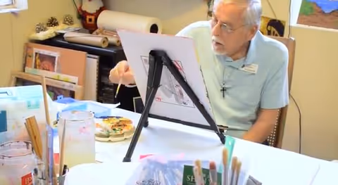 An elderly man wearing glasses and a light blue shirt is seated at a table with painting supplies, including brushes and a canvas on an easel. The setting appears to be an art or craft room with various art materials and paintings in the background.
