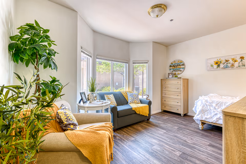 A bright and cozy living space with a beige armchair draped with an orange throw blanket and a patterned pillow, a gray loveseat with yellow and patterned pillows, and a small round white table between them holding a plant and picture frames. The room features large windows letting in natural light, a wooden chest of drawers, a bed with white bedding, a wall clock, and a sunflower-themed painting. There are several green plants adding a touch of nature to the room.