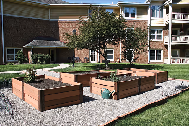 Raised garden beds with soil and some small plants in an outdoor area of a senior living facility. A green watering can is placed near one of the beds. In the background, there is a brick building with windows, a tree, and a pathway.