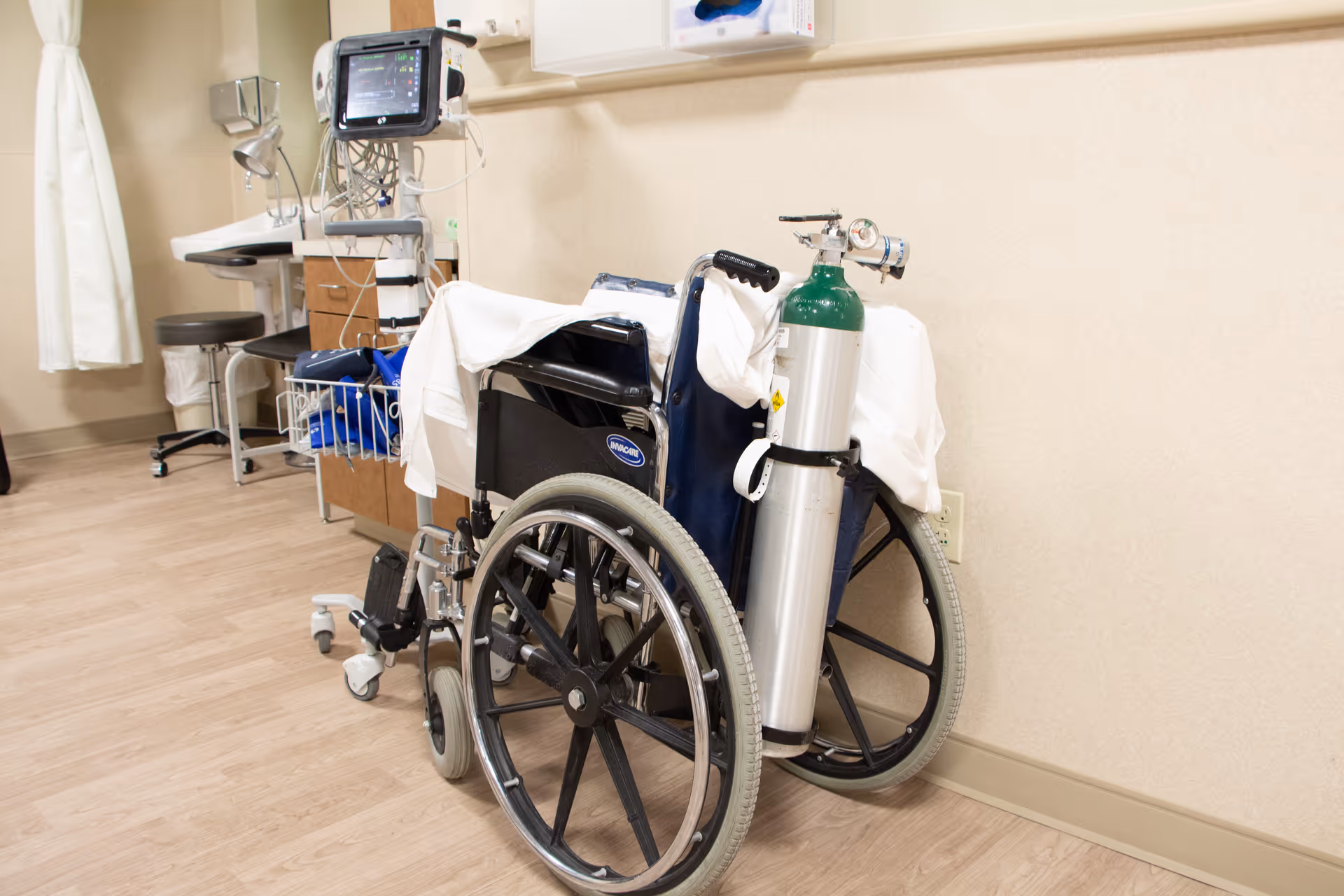 A wheelchair with an attached oxygen tank is positioned in a medical room with light wood flooring and beige walls. In the background, there is medical equipment including a monitor, a stool, and a small desk with drawers. A white curtain is partially visible on the left side.