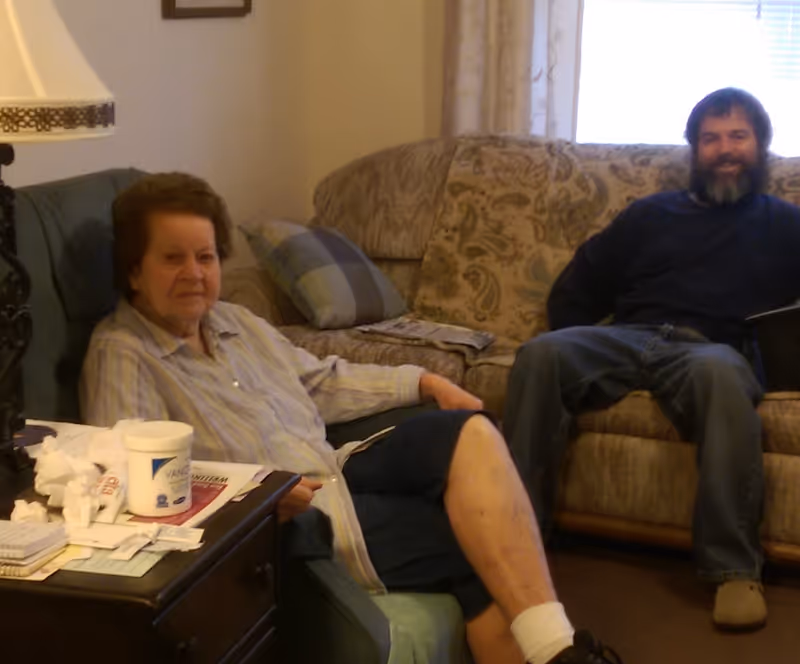 Two people seated on upholstered sofas in a cozy living room area with a side table and lamp.