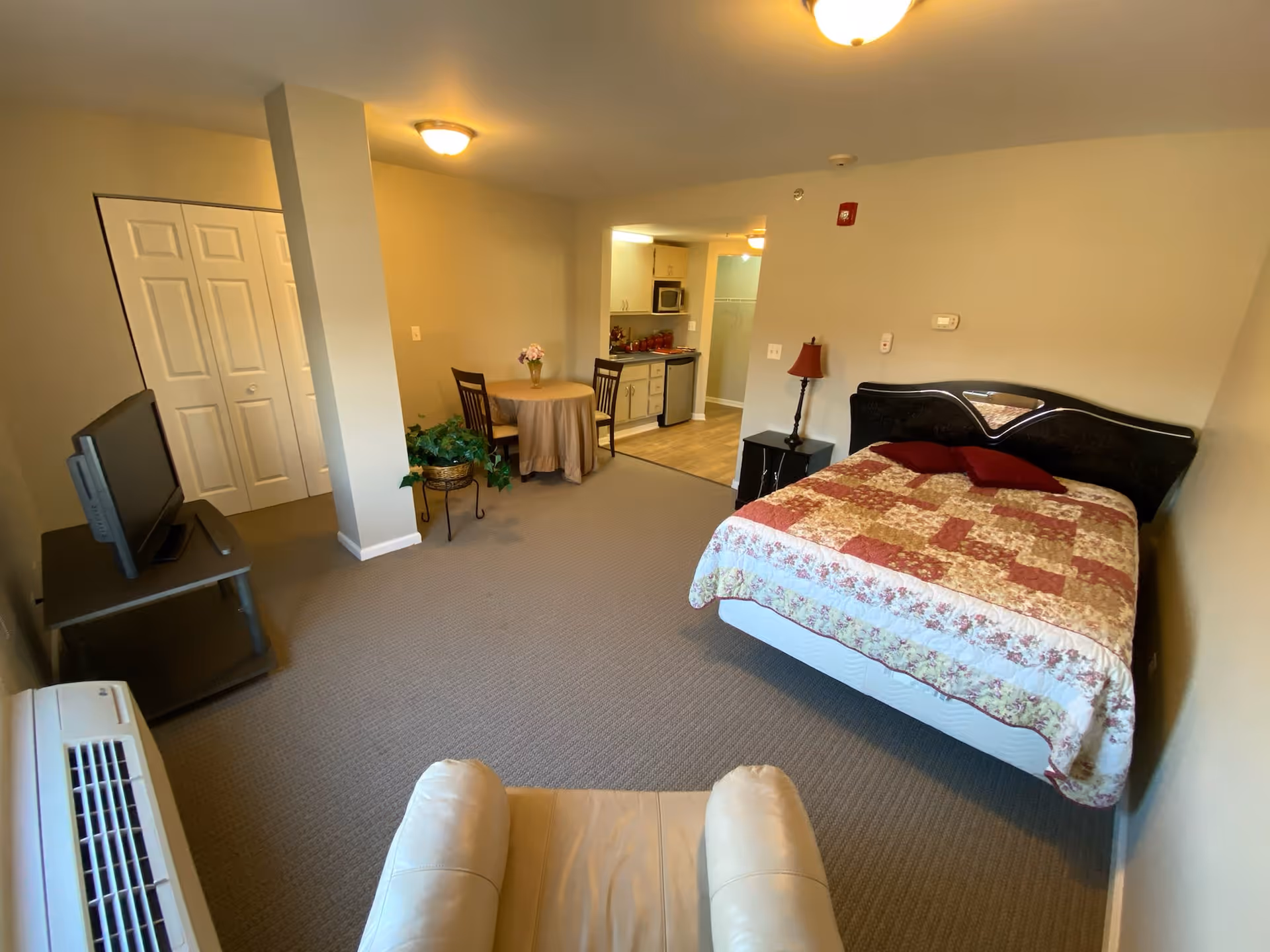 A spacious assisted living studio apartment featuring a bed with a patterned quilt, a small round dining table with two chairs, a kitchenette with cabinets, microwave, and mini fridge, a TV on a stand, a beige recliner chair, and a closet with double doors. The room has beige walls and carpeted flooring.
