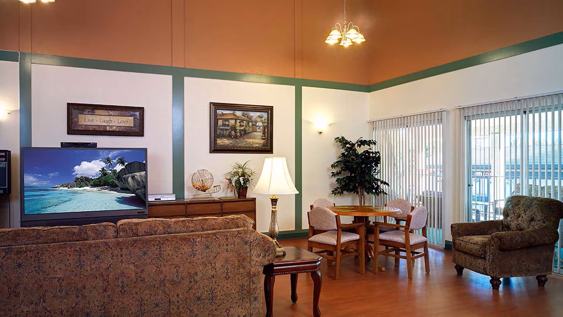 A cozy living room area with a patterned sofa facing a flat-screen TV displaying a beach scene. The room has a wooden floor, a round wooden table with four chairs, a single armchair, a floor lamp, framed artwork on the walls, and large windows with vertical blinds letting in natural light.