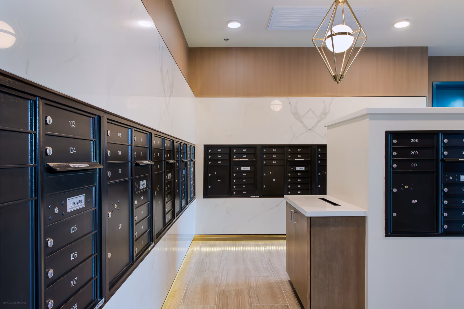Interior view of a modern mailroom with multiple black mailboxes mounted on white marble walls, a wooden cabinet with a white countertop and a mail slot, and a geometric pendant light hanging from the ceiling.