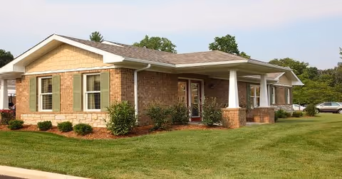 Single-story brick building with green shutters and a covered entrance, surrounded by a well-maintained lawn and some small bushes, with a few trees and parked cars in the background under a clear sky.