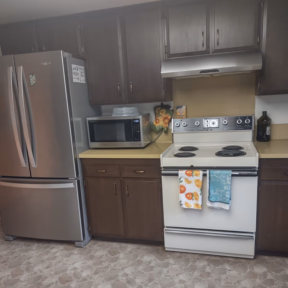 A kitchen area with dark brown cabinets, a stainless steel refrigerator, a microwave on the countertop, and a white electric stove with two colorful kitchen towels hanging on the oven door. There is a sunflower-themed oven mitt hanging on the wall and a bottle of cooking oil on the counter.