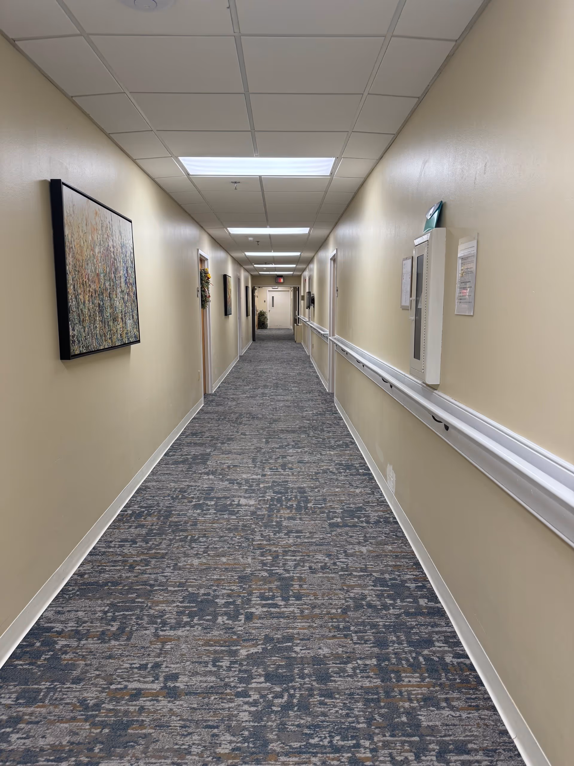 A long, well-lit hallway in a senior living facility with beige walls, patterned carpet flooring, handrails on both sides, several closed doors, framed artwork on the walls, and fluorescent ceiling lights.