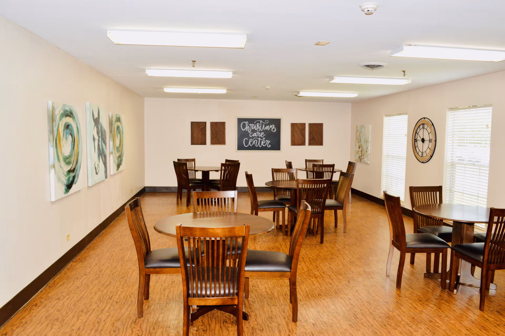 A dining room with several round wooden tables and matching chairs arranged neatly on a wood floor. The walls are light-colored and decorated with abstract paintings, a large clock, and a sign that reads 'Christian Care Center'. The room is well-lit with ceiling lights and has windows with blinds on one side.