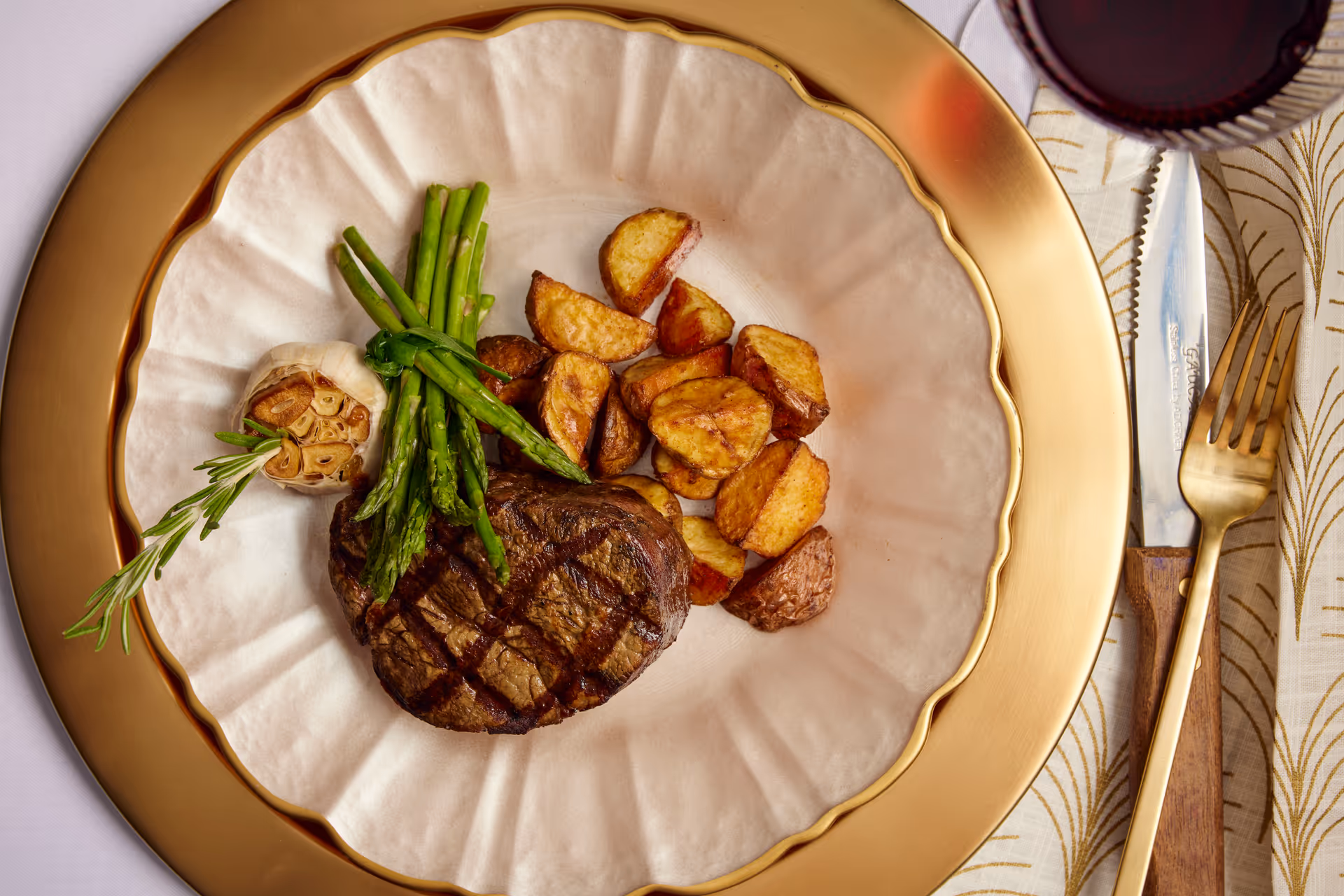 A plated meal consisting of a grilled steak topped with asparagus, roasted potato wedges, a whole roasted garlic bulb, and a sprig of rosemary on a white plate with a gold rim. The plate is set on a gold charger with a knife and fork on a patterned napkin, and a glass of red wine is partially visible in the top right corner.