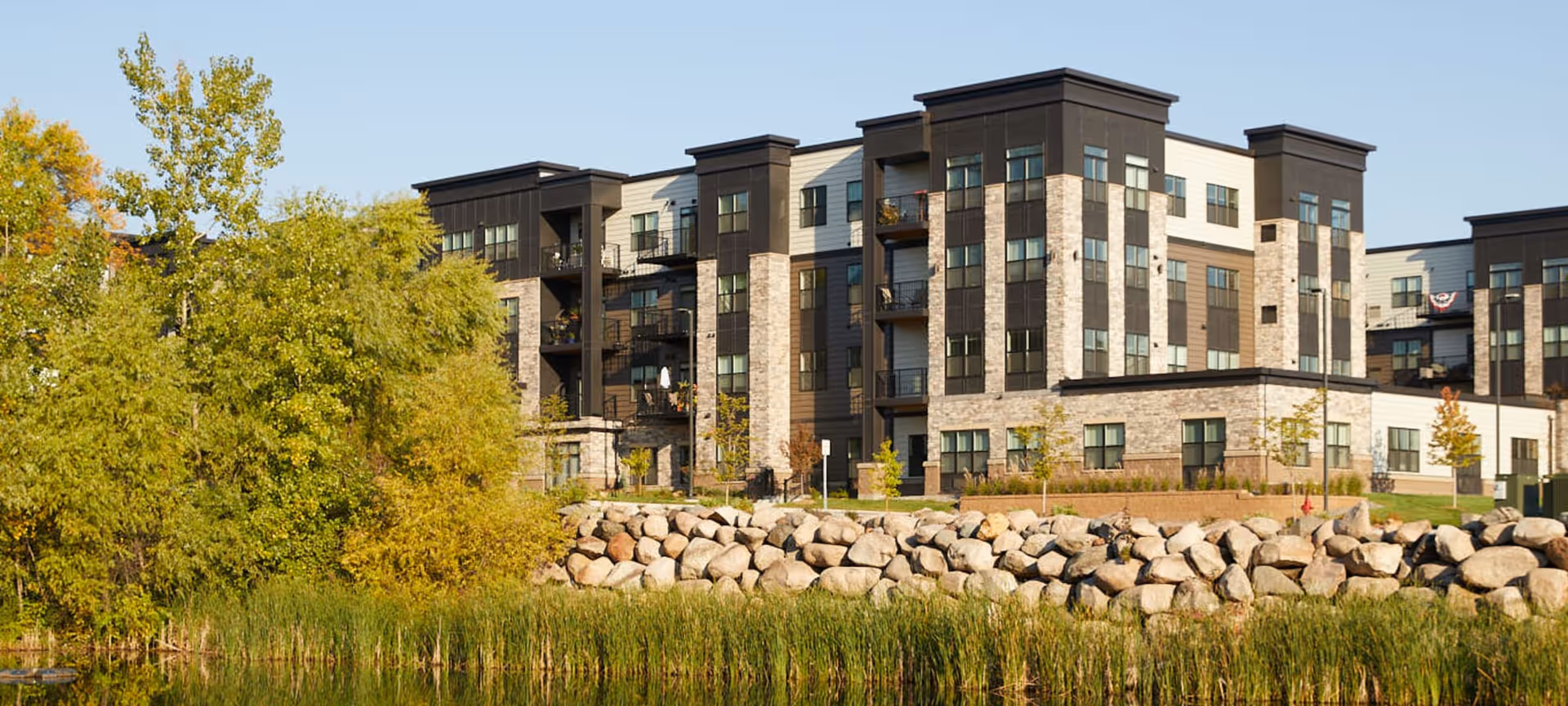 Exterior view of a modern multi-story residential building with balconies, surrounded by trees and a pond with tall grass and rocks in the foreground under a clear sky.