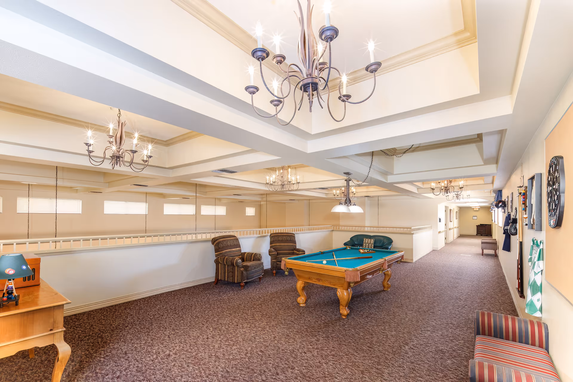 A spacious indoor recreational area with a pool table in the center, surrounded by comfortable chairs and a small couch. The room features multiple chandeliers hanging from a coffered ceiling, carpeted floors, and a long hallway extending to the right. There is a dartboard and other game accessories mounted on the wall.