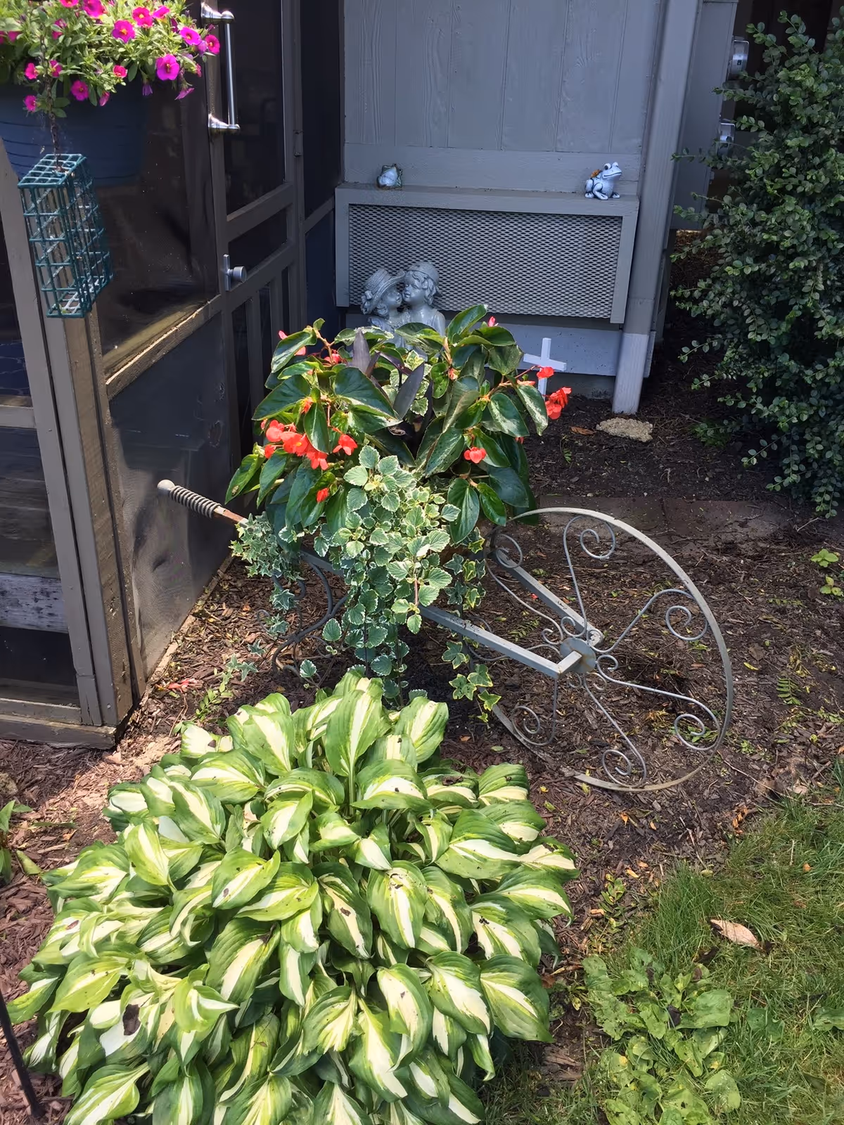 A garden area with various green plants and flowers, including a large leafy plant in the foreground and a decorative metal wheelbarrow planter with red flowers and ivy. There is a small statue of two children kissing near a wall with a vent, and a hanging basket with pink flowers on the left side near a screened door.