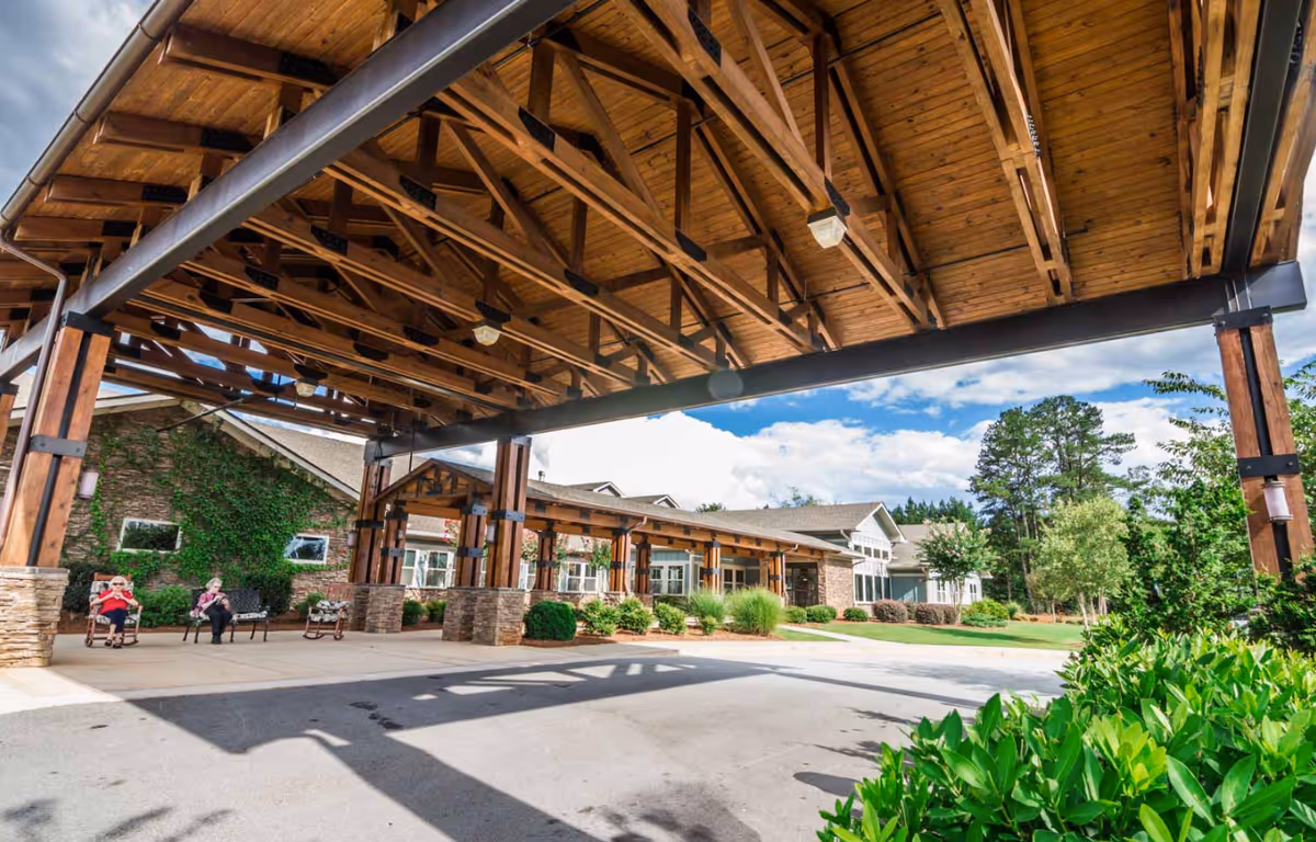 Covered wooden porte-cochere at the front entrance of a senior living building with benches and a few people seated nearby.