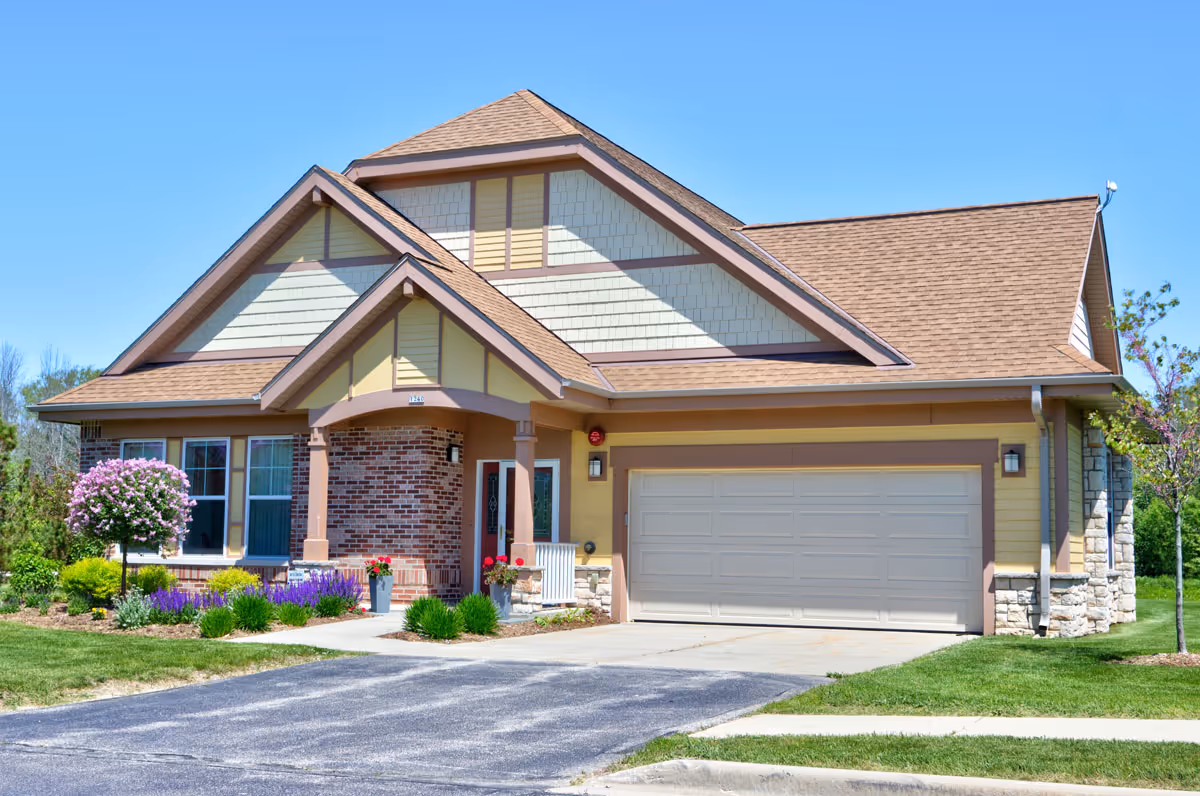 Exterior view of a single-story residential building with a gable roof, a two-car garage, and a small front porch. The house features a combination of brick, stone, and siding with beige and brown tones. There is a well-maintained lawn with shrubs, flowers, and a small tree in front of the house under a clear blue sky.