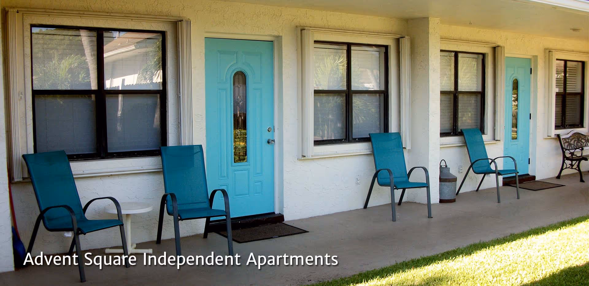 Outdoor patio area of Advent Square Senior Living with turquoise doors and matching turquoise chairs arranged along the wall under windows, with a small white side table and a bench at the far end.