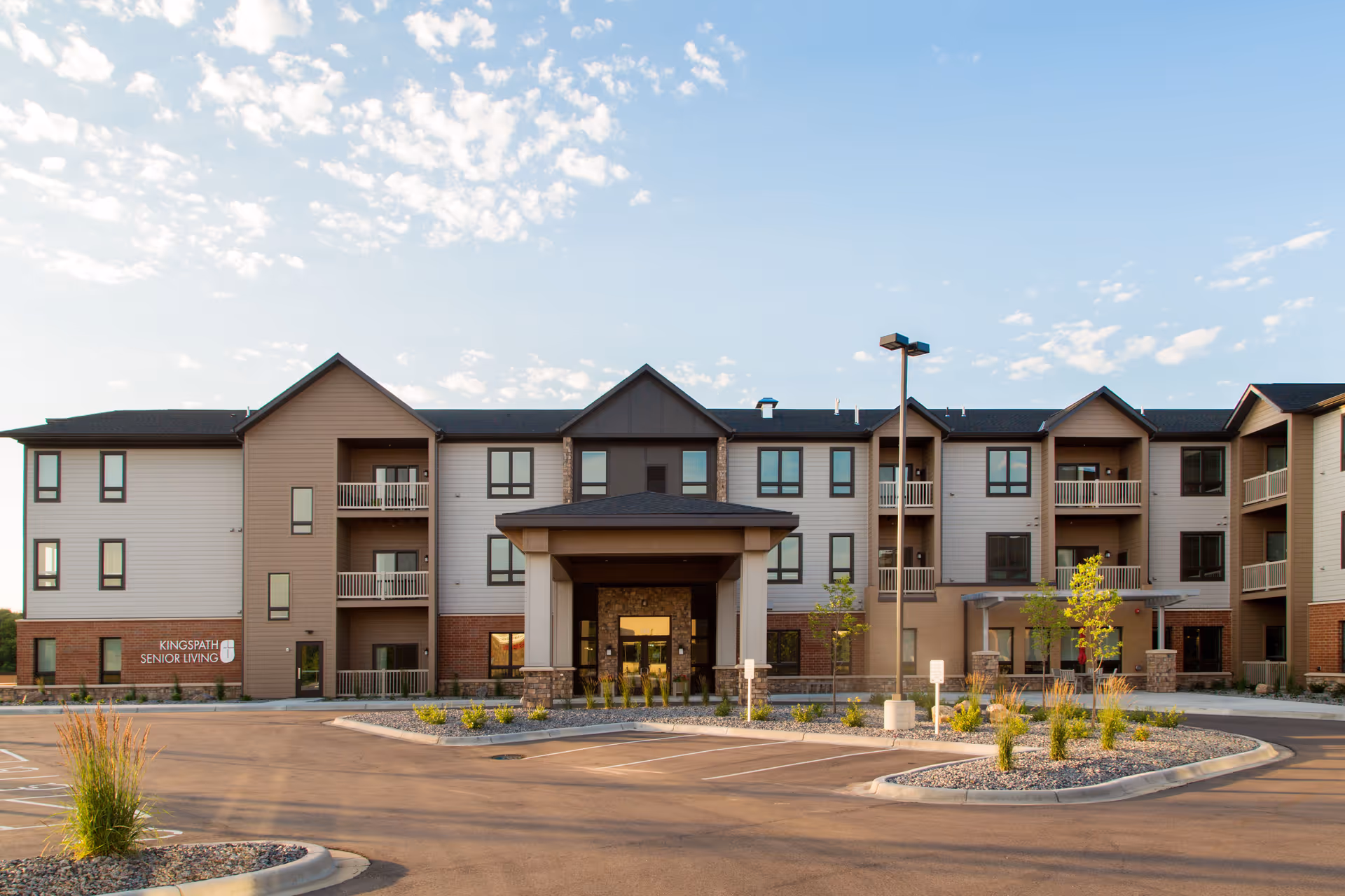 Exterior front view of a three-story senior living facility building with balconies, a covered entrance, and a parking lot in front under a partly cloudy sky.