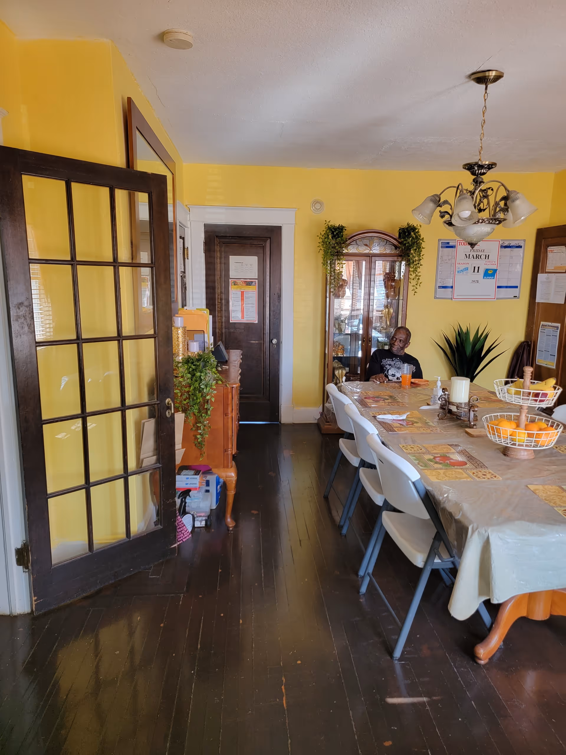 A dining room with yellow walls and dark wooden floor. There is a long table covered with a tablecloth, surrounded by white folding chairs. On the table are placemats, a tiered fruit basket with oranges and bananas, and a candle centerpiece. A man is sitting at the far end of the table. The room has a glass-paneled door on the left, a wooden cabinet with plants, and a wall calendar showing the date March 11. A chandelier with five lights hangs from the ceiling.
