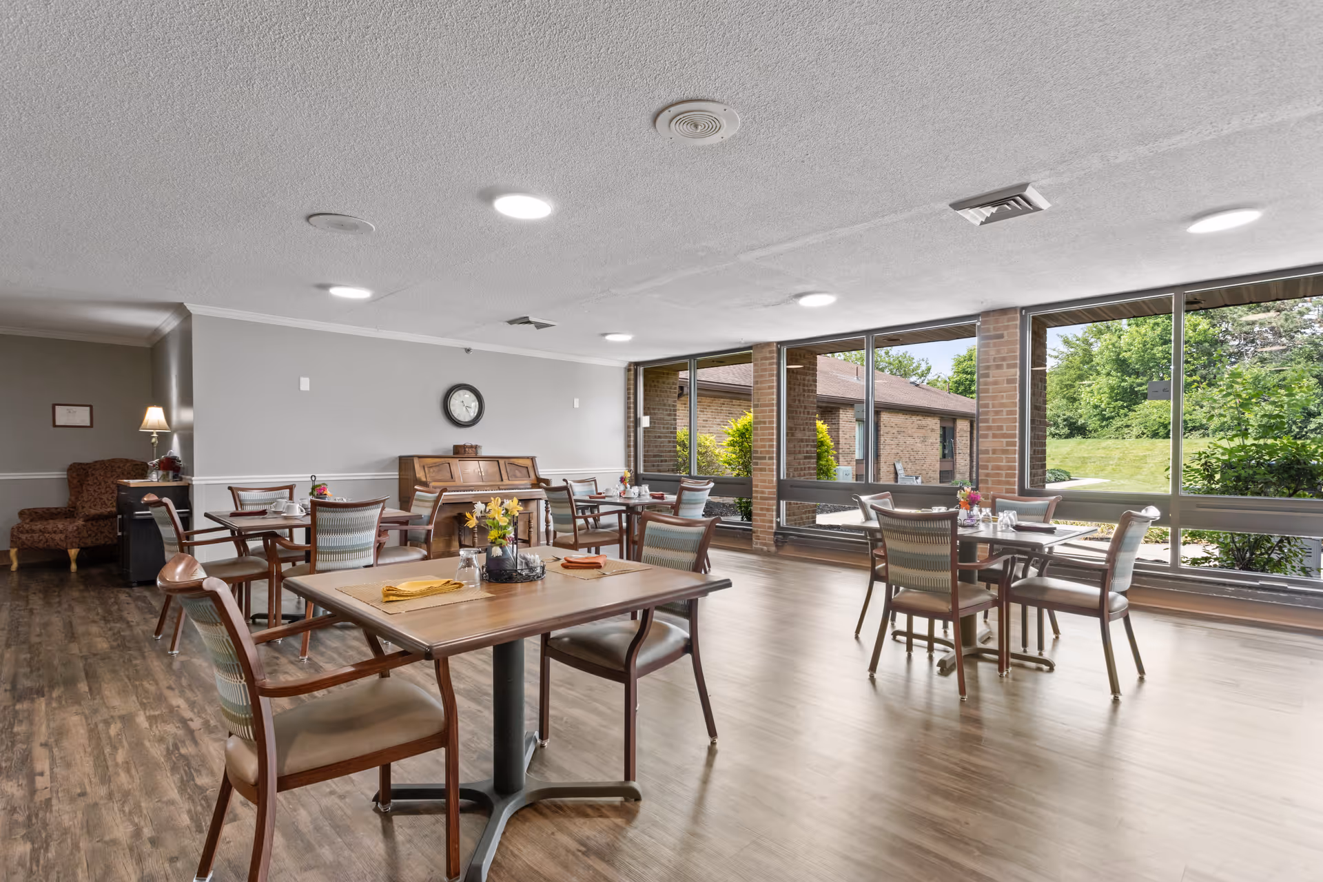 A bright dining room with several wooden tables and chairs arranged neatly. The room has large windows letting in natural light and offering a view of greenery outside. There is a piano against the far wall, a clock above it, and a cozy armchair with a lamp in the corner. The floor is wooden and the ceiling has recessed lighting.