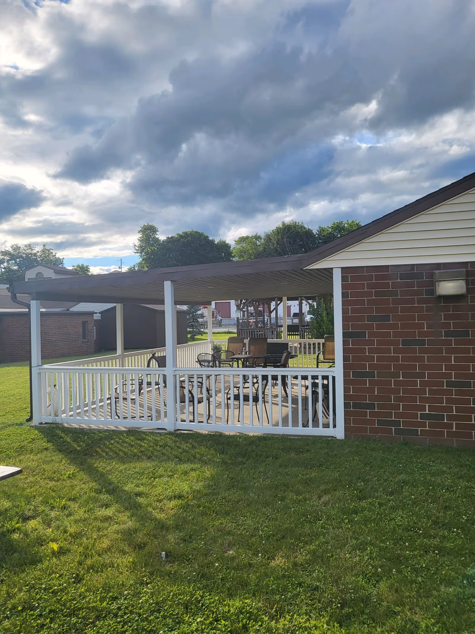 Outdoor covered patio area with a white railing and several chairs and tables underneath. The patio is attached to a brick building and surrounded by green grass. The sky above is partly cloudy.