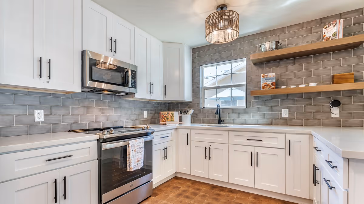 Modern kitchen with white cabinets, stainless steel microwave and oven, gray subway tile backsplash, wooden floating shelves with cookbooks, a window above the sink, and a ceiling light fixture.