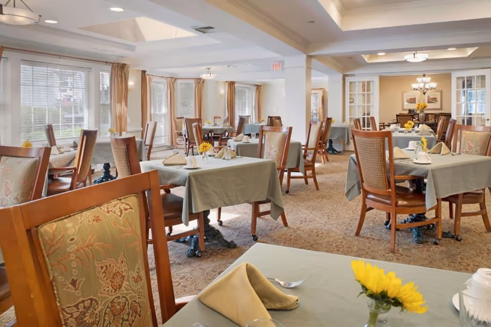 A bright and spacious dining room with multiple tables covered in green tablecloths, each set with napkins, cups, and small vases holding yellow flowers. The room features large windows with beige curtains allowing natural light to fill the space, wooden chairs with patterned cushions, and a carpeted floor. The ceiling has recessed lighting and a skylight, and there is a chandelier in the background.