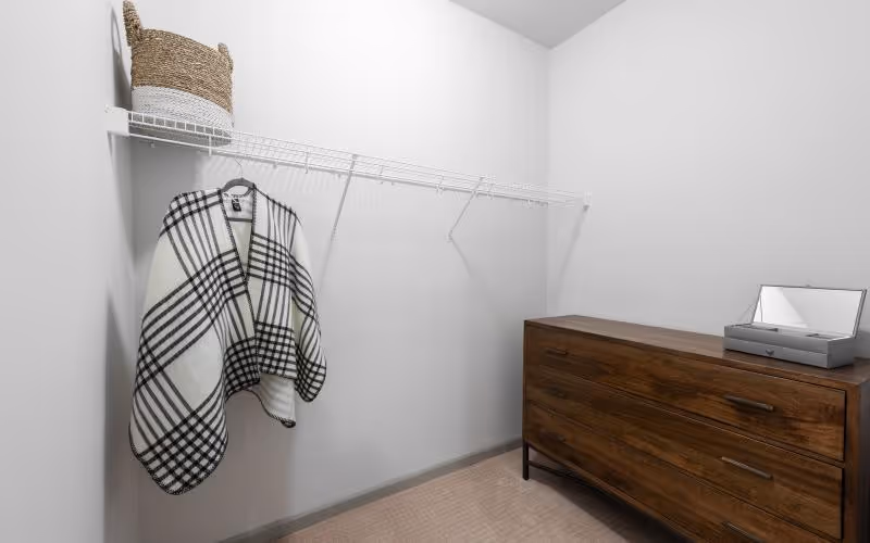 Walk-in closet with white wire shelving, a plaid garment hanging, a wicker basket on the shelf, and a wooden dresser with an open jewelry box.