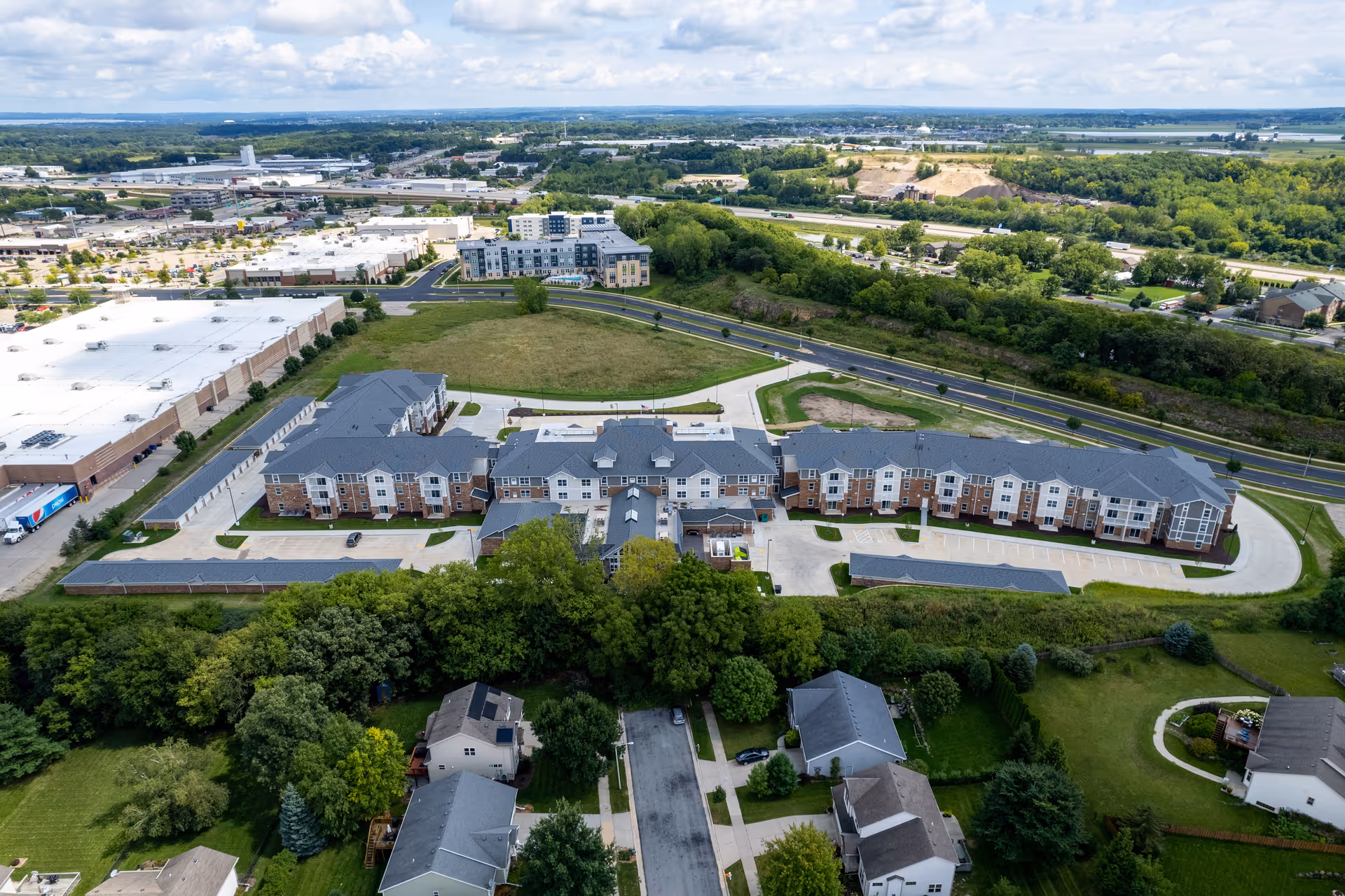 Aerial view of a multi-wing retirement community building with parking lots, surrounding roads, green spaces, and neighboring houses.