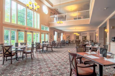 A spacious dining room with multiple wooden tables and chairs arranged neatly. The room features large windows allowing natural light to fill the space, high ceilings with chandeliers, and a balcony overlooking the area. The carpet has a patterned design, and the walls are painted in warm tones.