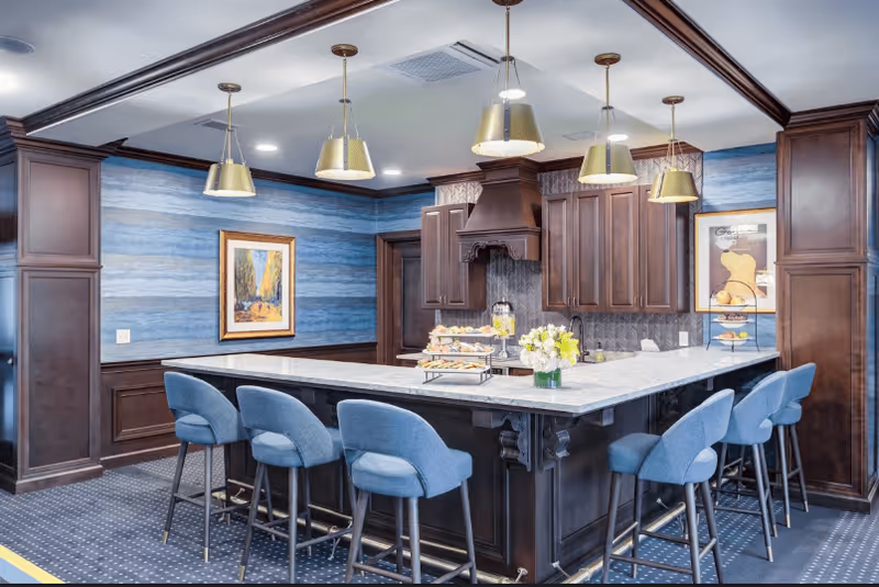 Community kitchen with a large marble-topped island, blue upholstered barstools, pendant lights, and dark wood cabinetry.